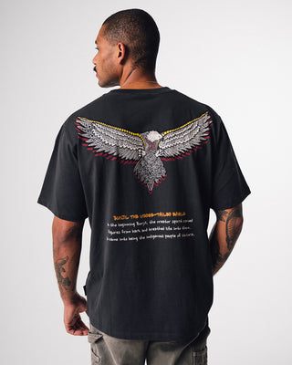 bunjil the wedged-tailed eagle indigenous art on the back of a t-shirt in a washed out black color  being worn. Text on t-shirt below art says 'in the beginning bunjil, the creator spirit carved figures from bark and breathed life into them. so came into being the indigenous people of Victoria.'