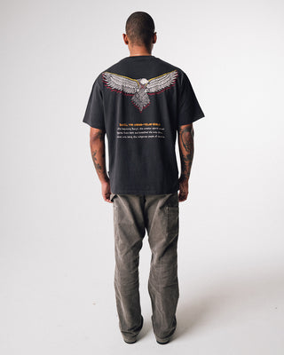 bunjil the wedged-tailed eagle indigenous art on the back of a t-shirt in a washed out black color being worn in a full length image. Text on t-shirt below art says 'in the beginning bunjil, the creator spirit carved figures from bark and breathed life into them. so came into being the indigenous people of Victoria.'