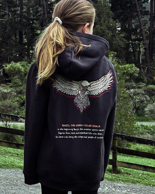 bunjil the wedged-tailed eagle indigenous art on the back of a hoodie in a washed out black color being worn. Text on hoodie below art says 'in the beginning bunjil, the creator spirit carved figures from bark and breathed life into them. so came into being the indigenous people of Victoria.' image is shot in the forest with large trees and bushes