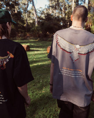 bunjil the wedged-tailed eagle indigenous art on the back of a t-shirt being worn. Text on t-shirt below art says 'in the beginning bunjil, the creator spirit carved figures from bark and breathed life into them. so came into being the indigenous people of Victoria.' background is a outback forest with large trees and bright clear sky