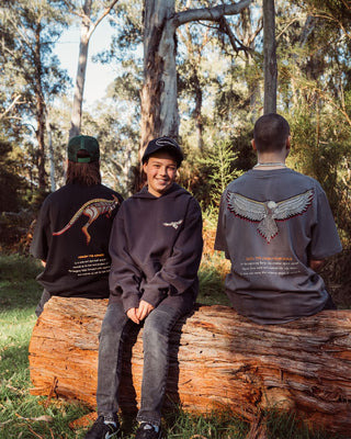 bunjil the wedged-tailed eagle indigenous art on the back of a t-shirt being worn. Text on t-shirt below art says 'in the beginning bunjil, the creator spirit carved figures from bark and breathed life into them. so came into being the indigenous people of Victoria.' background is a outback forest with large trees and bright clear sky. in the image there 3 kids sitting on a log all wearing murrundindi clothing