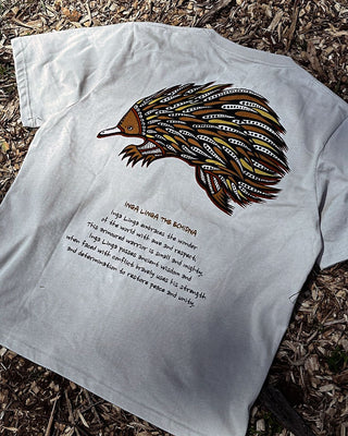 inga linga the echidna indigenous art on the back of a t-shirt in off white color in a detailed shot showing the full art. Text on t-shirt below art says 'Inga Linga embraces the wonder of the world with awe and respect. This armored warrior is small and mighty. Inga Linga possesses ancient wisdom and when faced with conflict bravely ueses his strength and determination to restore peace and unity.'