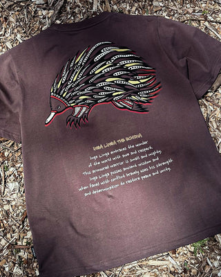 inga linga the echidna indigenous art on the back of a t-shirt in washed chocolate color in a detailed shot showing the full art. Text on t-shirt below art says 'Inga Linga embraces the wonder of the world with awe and respect. This armored warrior is small and mighty. Inga Linga possesses ancient wisdom and when faced with conflict bravely ueses his strength and determination to restore peace and unity.'